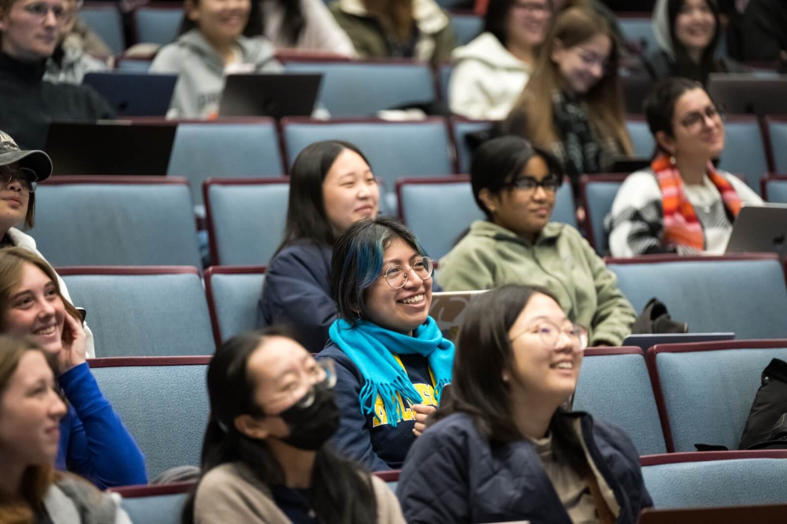 A group of diverse students sits in a lecture hall, attentively listening and smiling. Some students have laptops open on their desks. The atmosphere appears engaging and positive, with students seated in rows of light blue chairs.