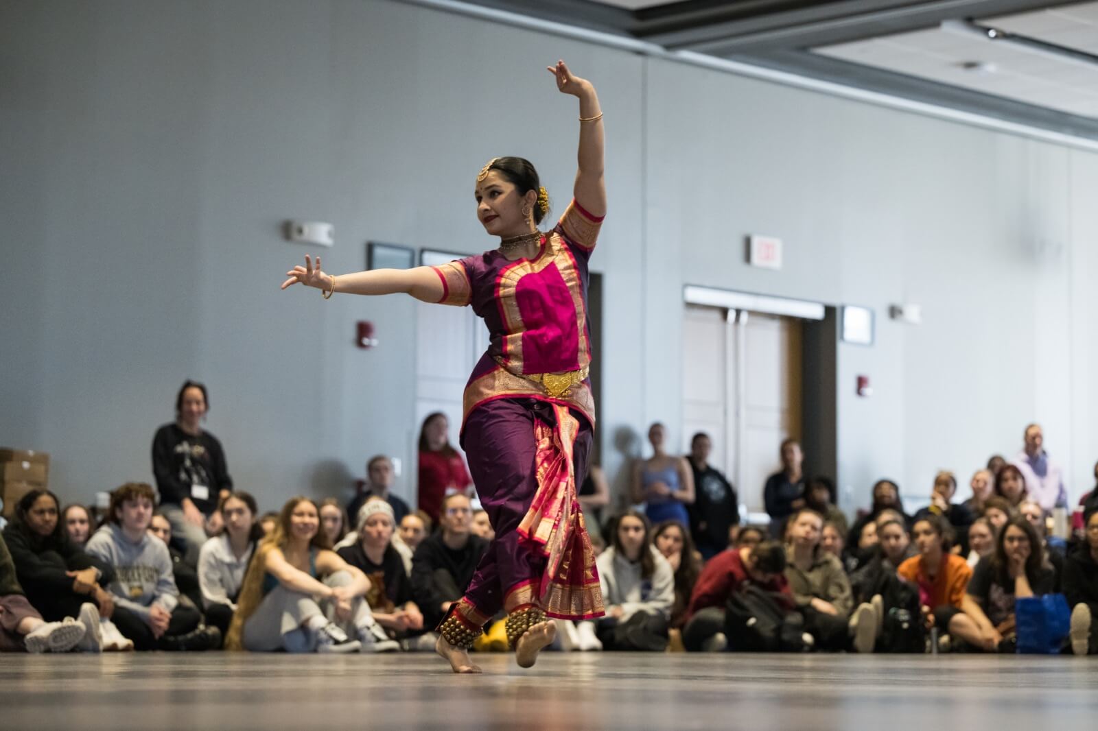 A dancer performs a traditional dance in vibrant clothing, including a pink and purple outfit with gold accents, amid an attentive audience seated around a spacious room. The dancer's graceful pose features one arm raised and the other extended to the side.