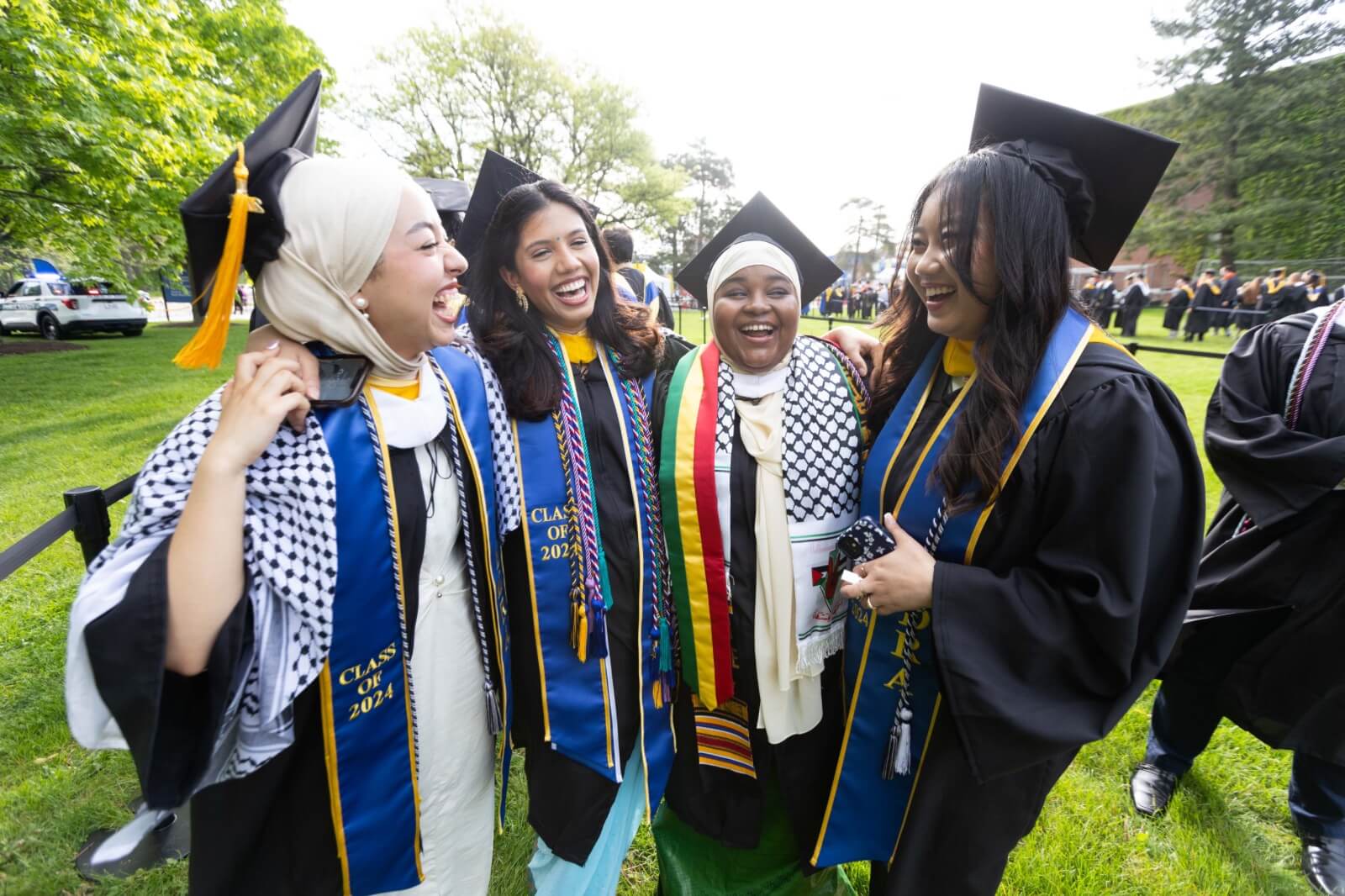 Four graduates in caps and gowns stand together, smiling and laughing. They wear blue and gold stoles with 