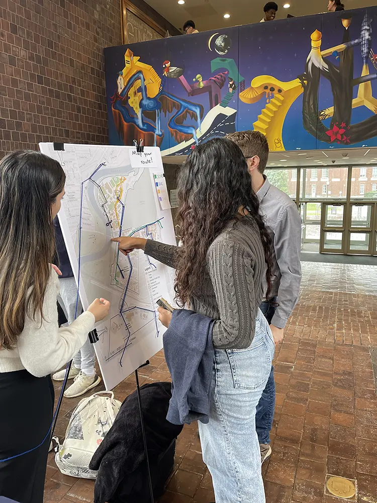 Three students map their travel routes with colored strings on a campus map displayed in Wilson Commons.