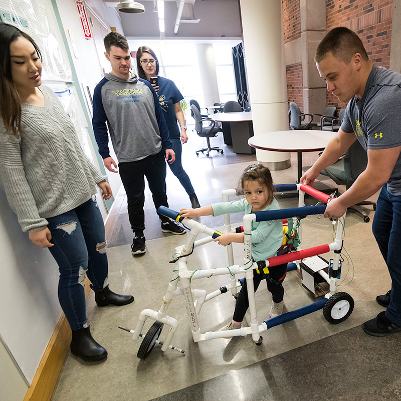 four students watch a child walk in their walker prototype, with wheels and handlebars and plastic piping