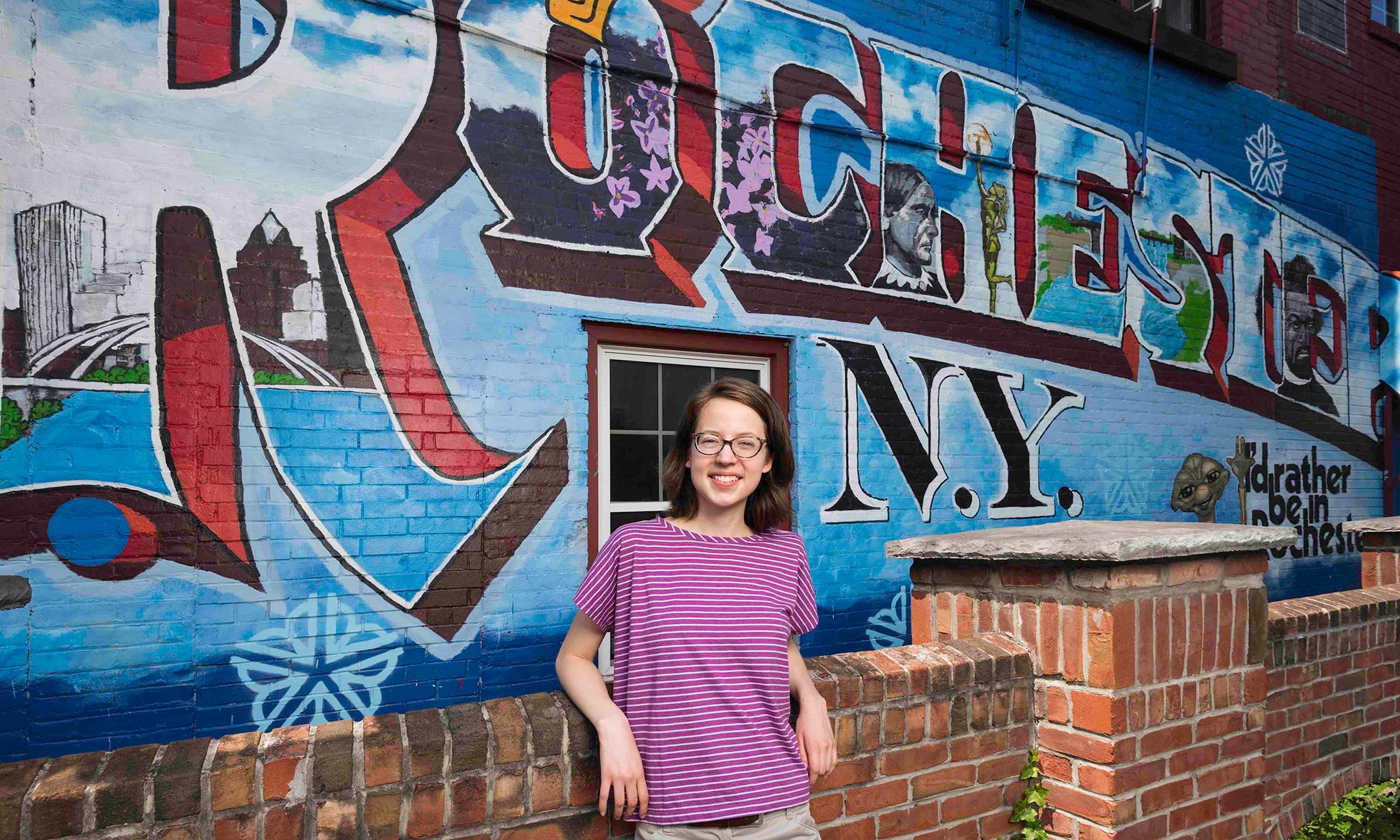 student stands in front of wall mural that reads ROCHESTER NY