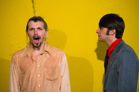 Two actors in front of a yellow backdrop being splashed with water. 