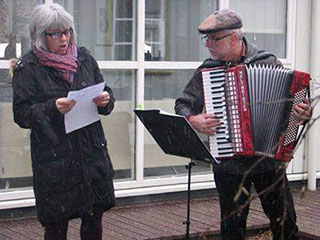 man playing accordian outside next to woman reading