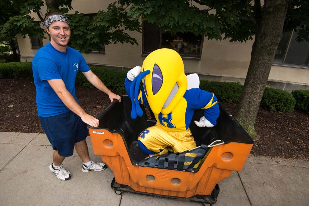 student pushes Rocky yellowjacket mascot in a luggage cart