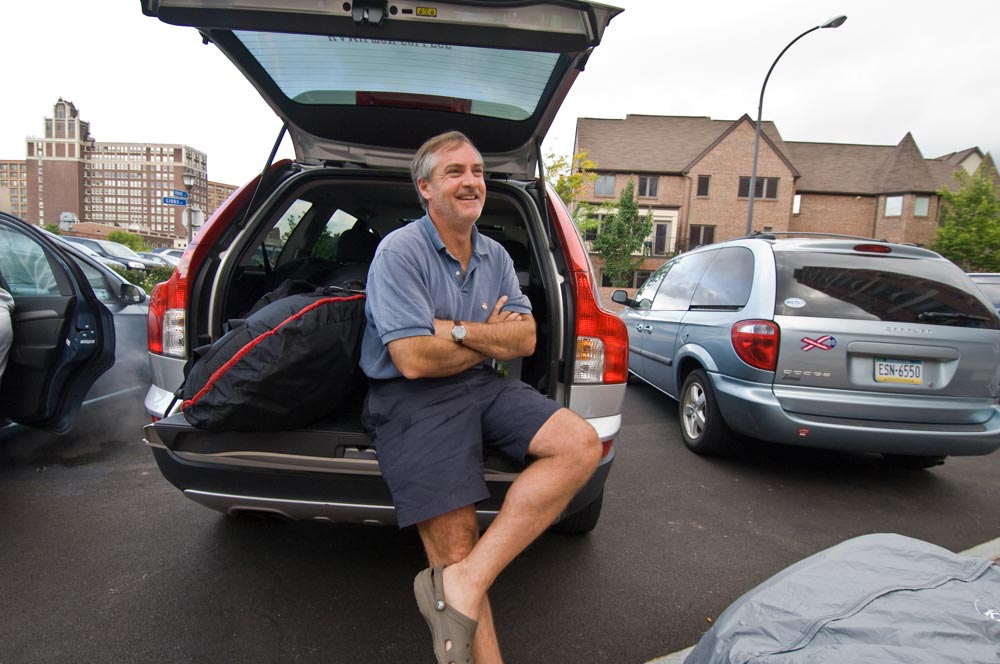 father sits in the bumper of his packed car and smiles