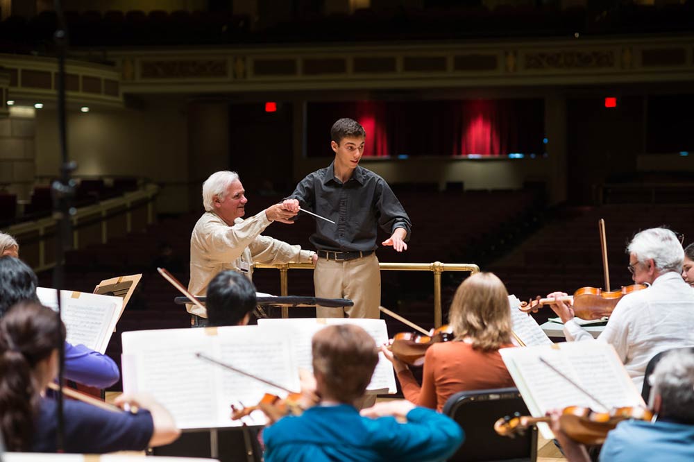 instructor helping a conductor at the front of an orchestra