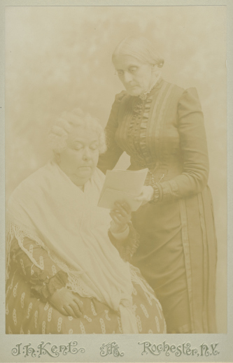 Seated Elizabeth Cady Stanton holds a document as Susan B. Anthony stands next to her.