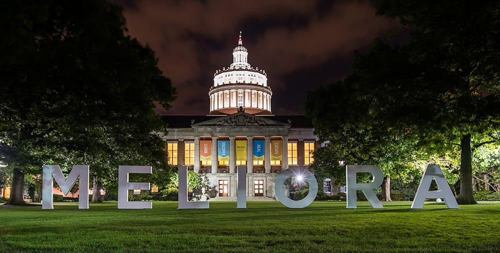 giant MELIORA letters lit up against Rush Rhees Library at night