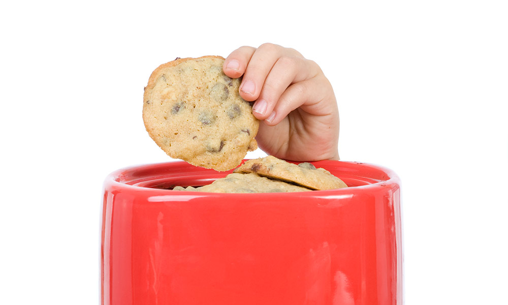 A child's hand reaches and draws a cookie from a jar.