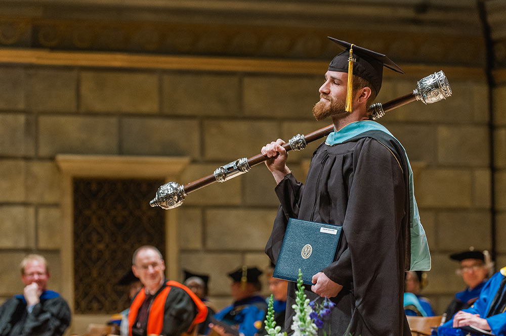 man in graduation robes carries the mace over his shoulder in Kodak Hall