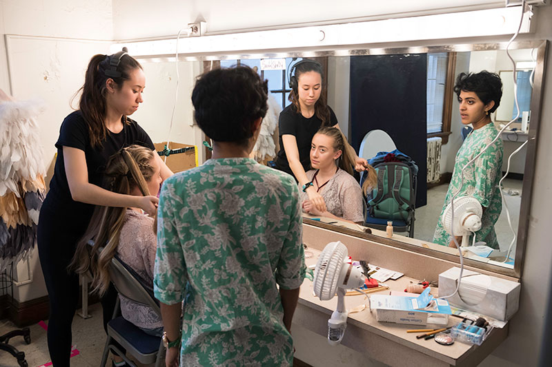 three students backstage, one getting make-up applied in front of a larger mirror