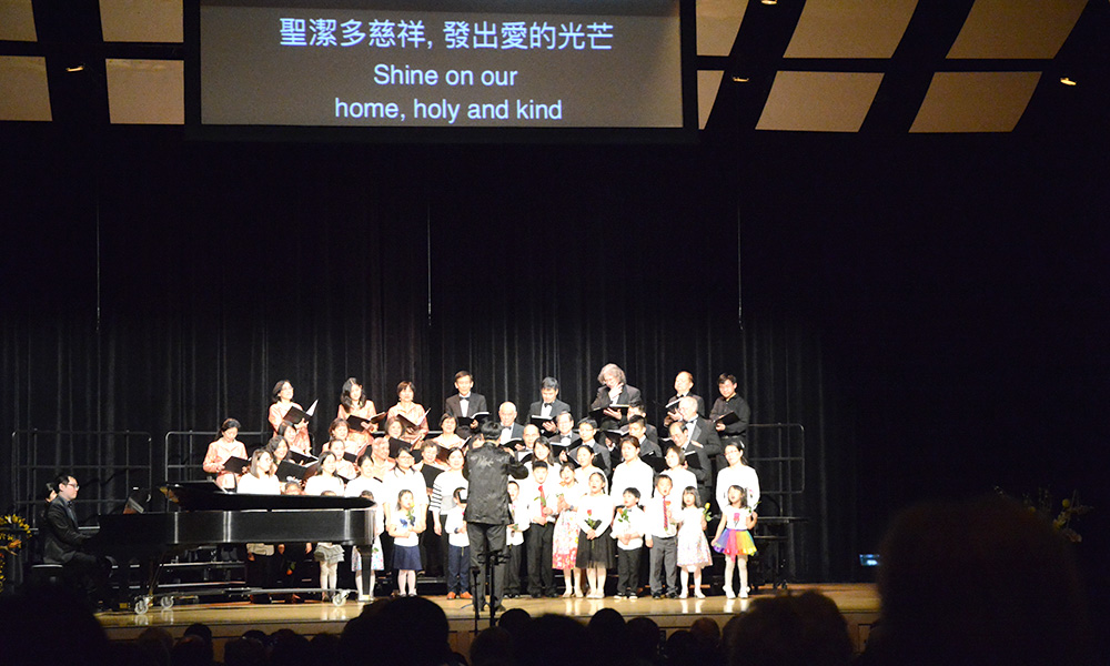 choir performing on stage, with screen above showing lyrics SHINE ON OUR HOME, HOLY AND KIND in English and Chinese