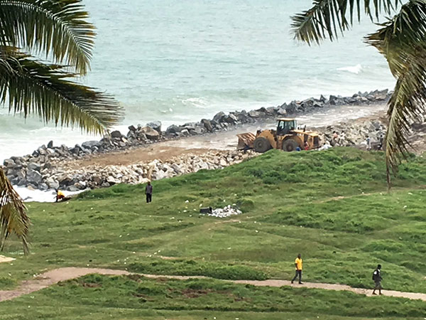 construction machine next to a stone wall on a beach as people walk past. 