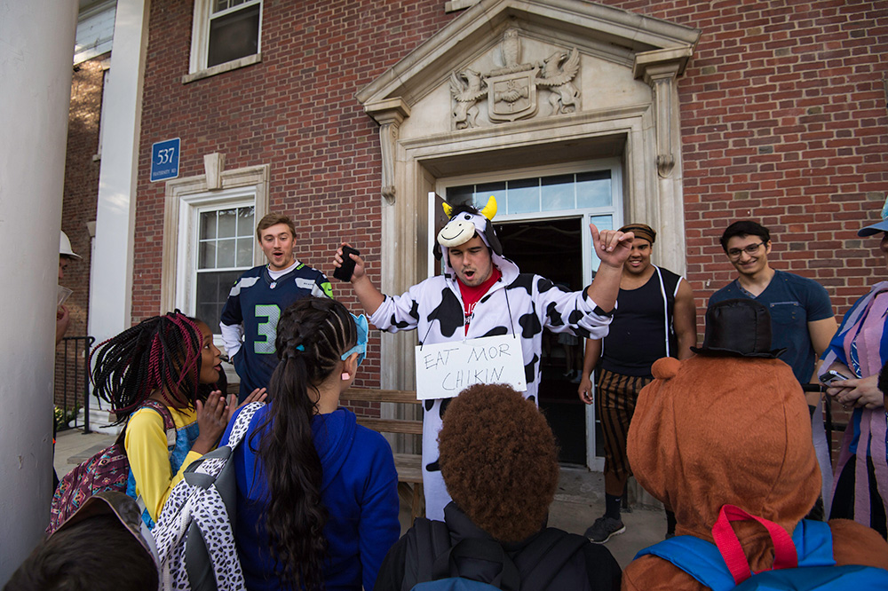 student dressed in cow costume speaks and laughs with a group of other students in front of a fraternity house