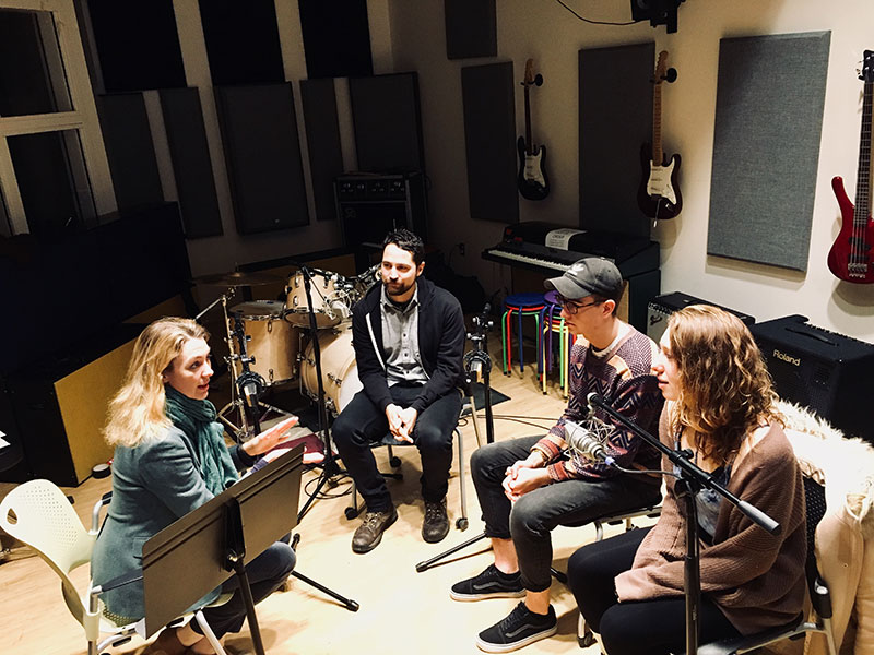 Four people sitting around microphones in a recording studio