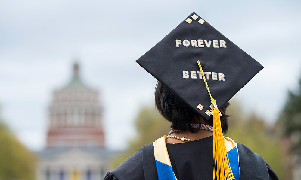 student in a cap and gown faces away from the camera and toward Rush Rhees Library; their graduation cap is decorated with the words FOREVER BETTER.