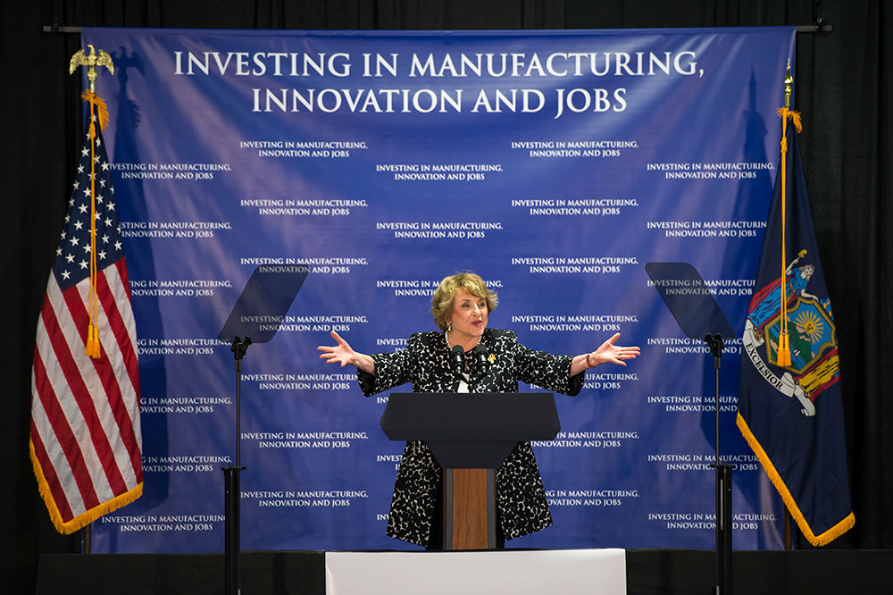 Louise Slaughter standing in front a large sign that reads INVESTING IN MANUFACTURING, INNOVATION AND JOBS