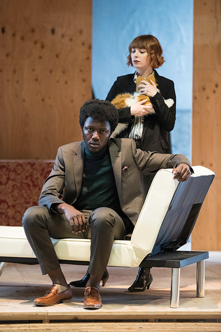 two actors on stage, one sitting on a couch and the other holding a cat