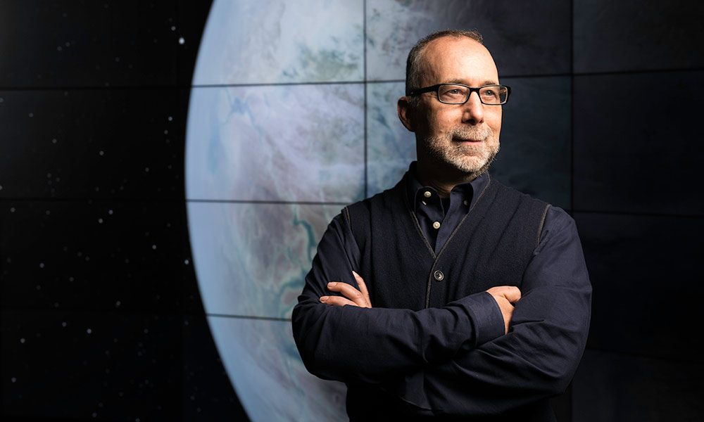 Adam Frank stands with arms crossed in front of a screen with a large photo of the Earth