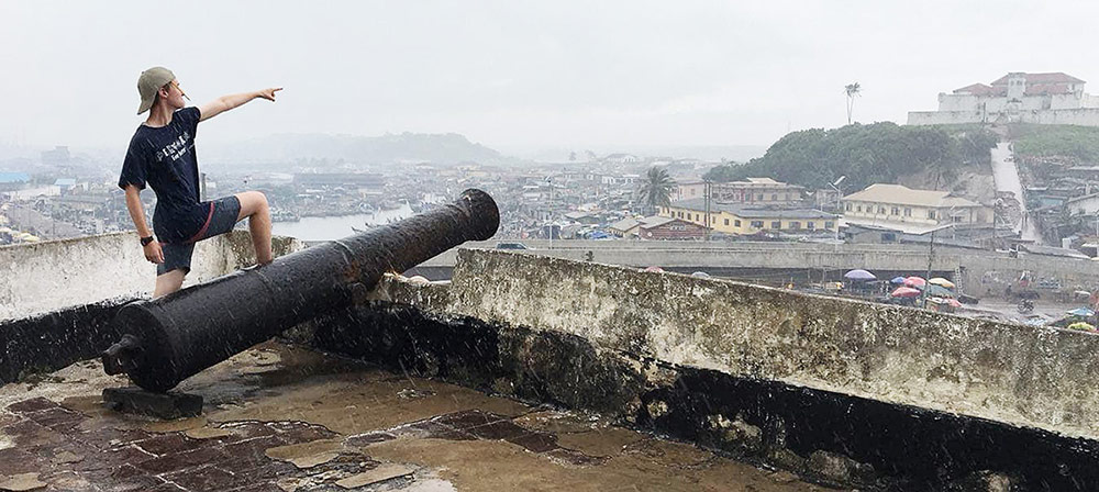 Ewan Shannon stands by a cannon, pointing to the town of Elmina from the rooftop of Fort Amsterdam