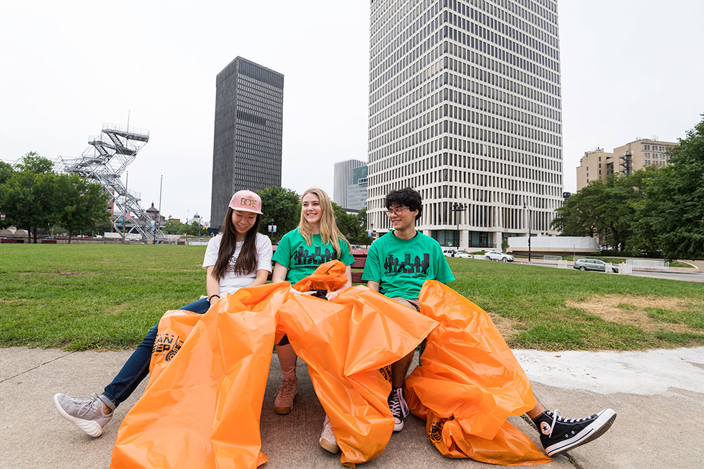 three students sitting on a park bench, relaxing and laughing, holding large garbage bags afer cleaning up a park