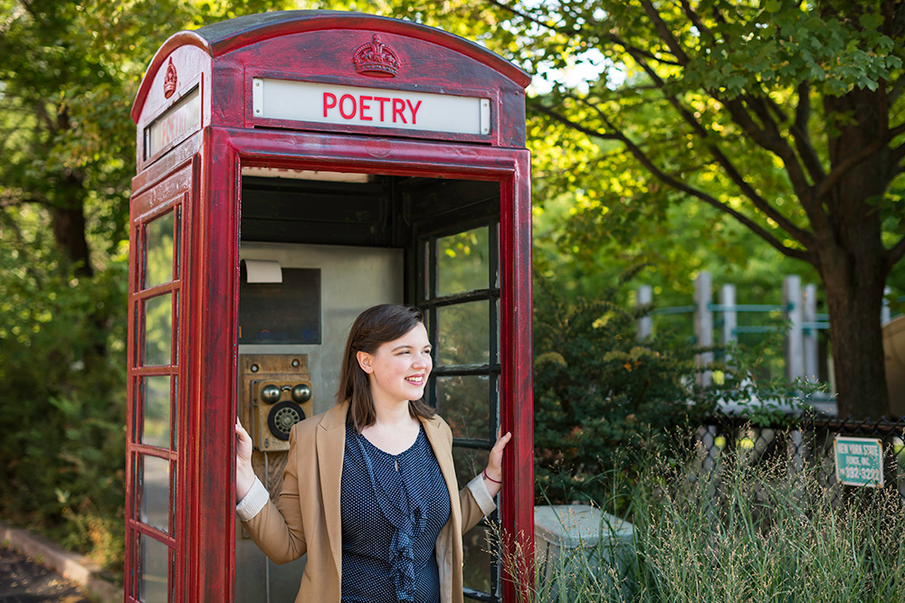Emily Tworek poses in an English style phonebooth that has the word POETRY written where the word TELEPHONE would be