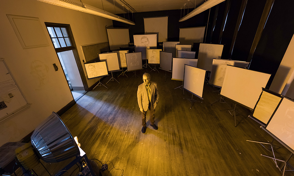 art professor stands in a disused classroom that has been filled with old-fashioned fimlstrip screens, each showing an image from an old high school yearbook.