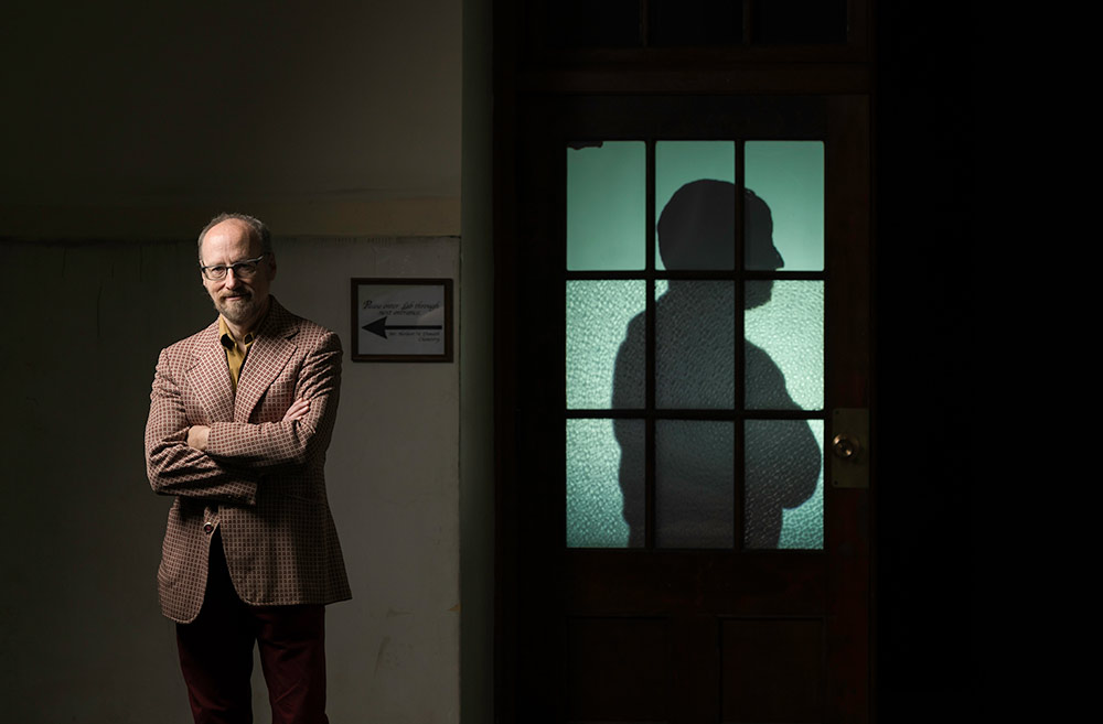 Alan Topolski stands with his arms crossed, and a silhoutte with its arms crossed is projected onto a classroom door next to him.