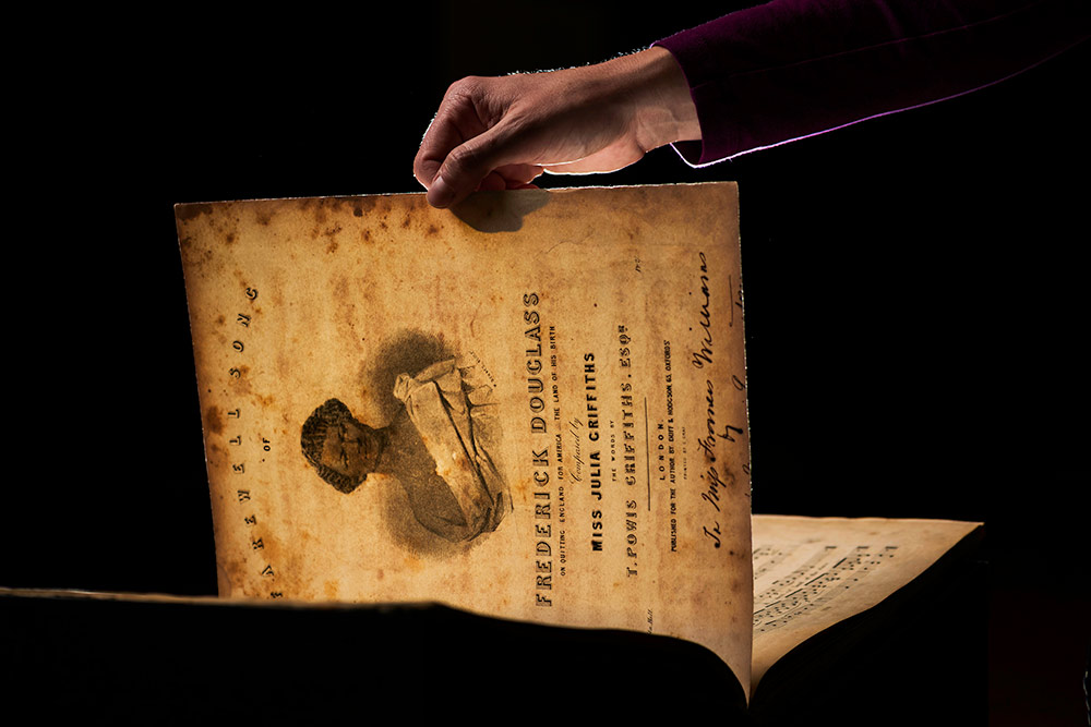 a hand holding up the page of a sheet music with Frederick Douglass's face.