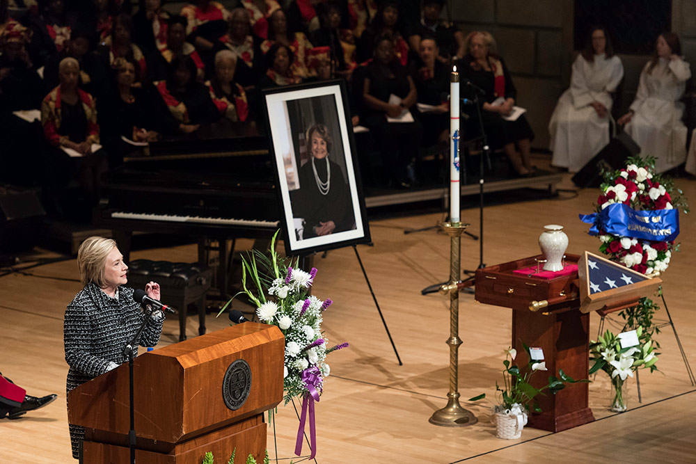 Hillary Clinton on stage at a funeral service with flowers and a portrait of Louise Slaughter.