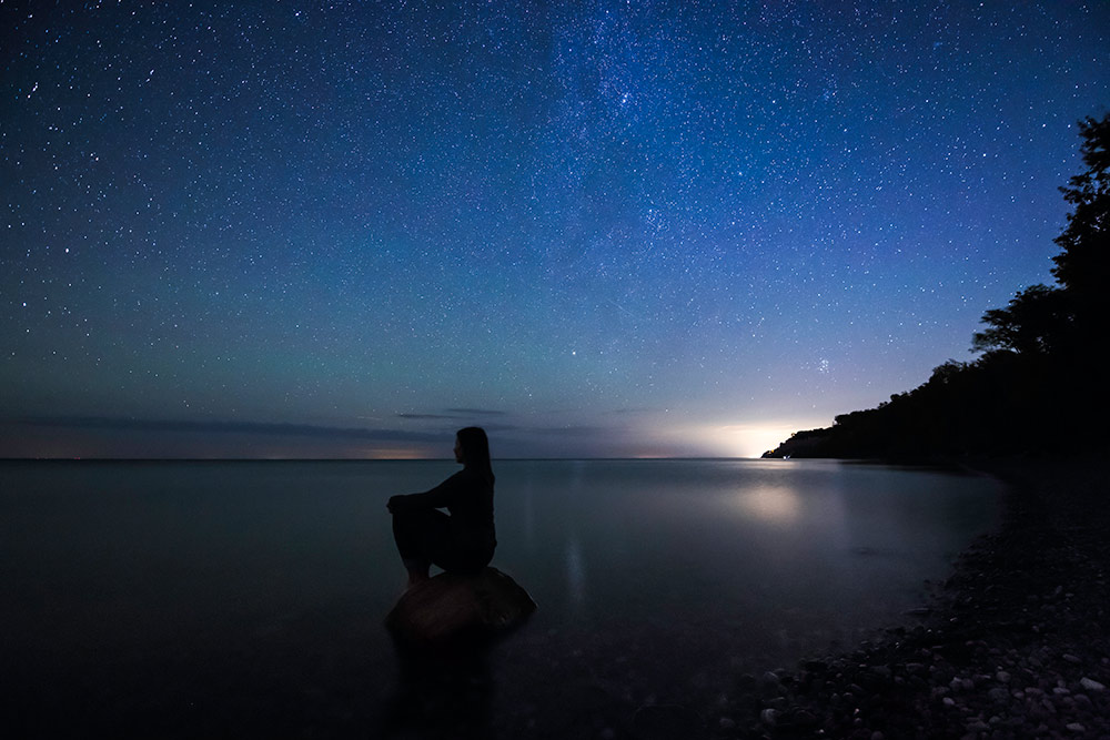 nighttime image of a person kneeling on a beach, with the lake and a sky full of stars in the background.