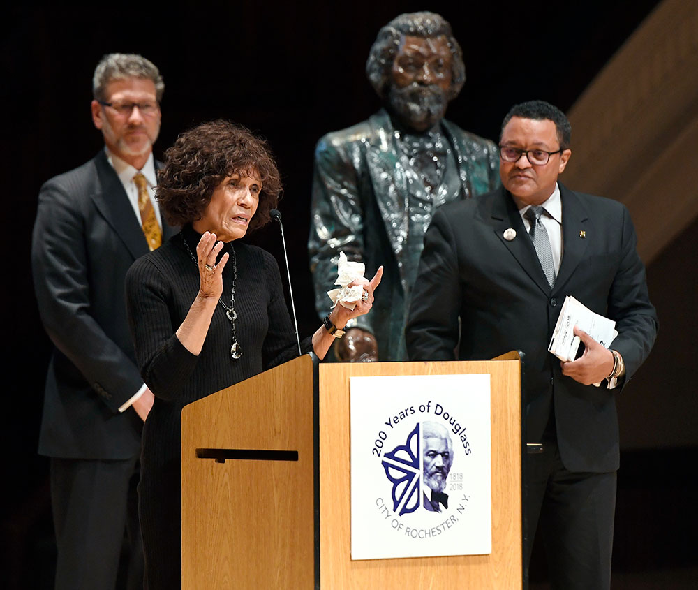 three people standing on stage, one behind a podium speaking, with a statue of Frederick Douglass in the background