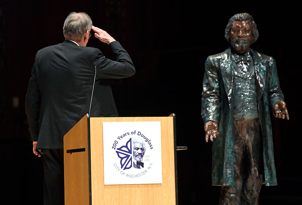 David Blight sands behind podium, turning to salute a statue of Frederick Douglass behind him. 