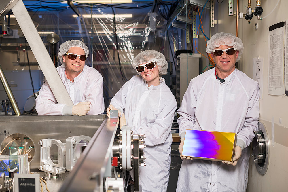 three researchers in clean suits and goggles, the one in the center holding a small, square prism-like device, and the one on the right holding a much larger version of the same device