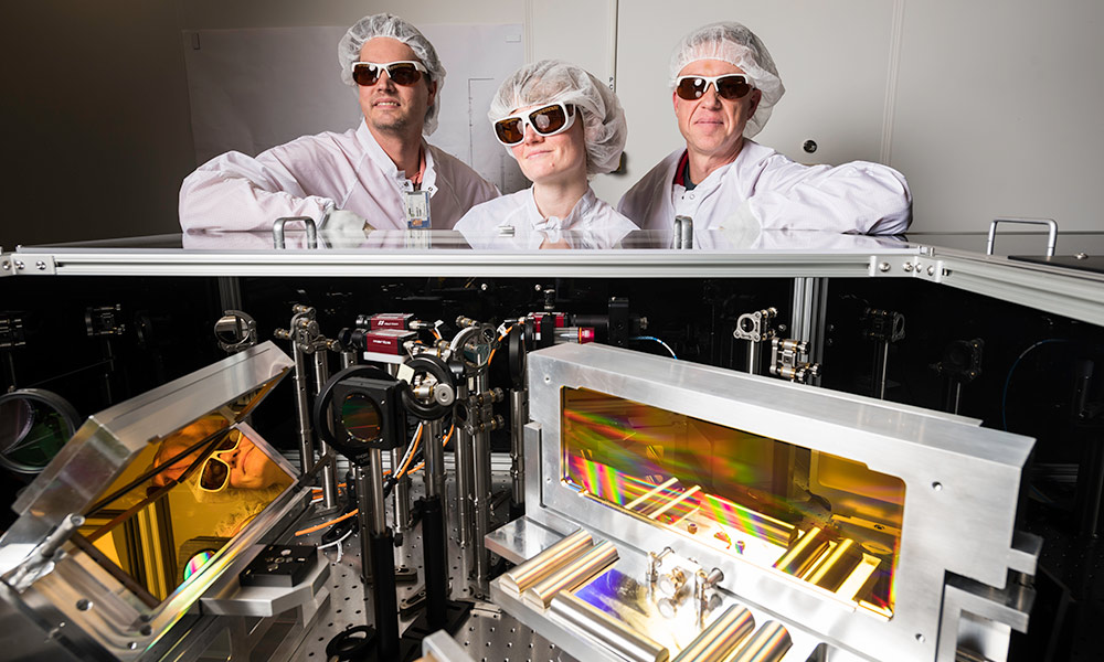three researchers wearing laser goggles and clean suits stand over an array of optical devices