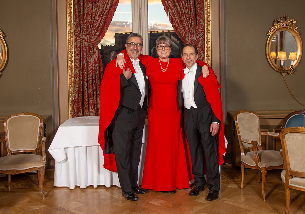 three people in formal attire, two in white tie and one in a red gown, smile and pose for a photo.