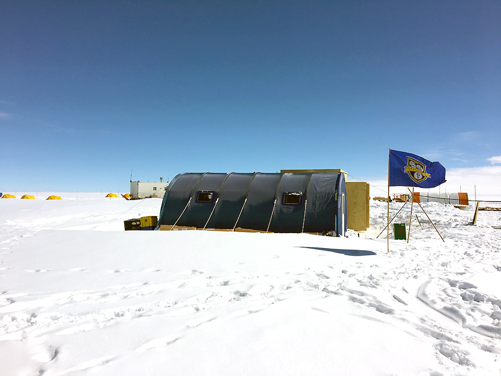 A blue tent sits on a field of ice and snow, with a University of Rochester MELIORA flag flying in front.