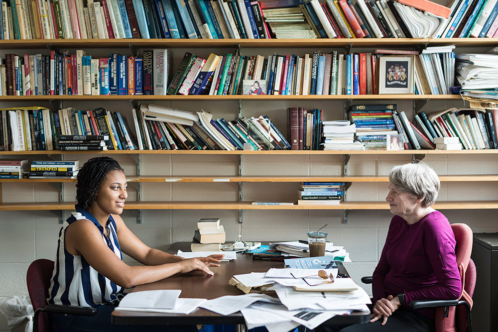 student and professor sit at an office table, the wall behind the filled with bookshelves