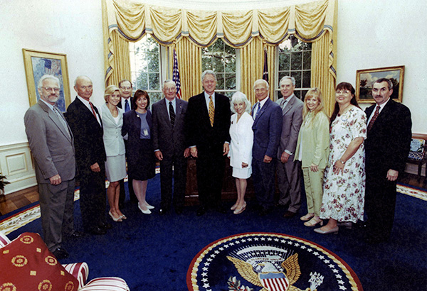 large group of people pose for a photo in the Oval Office, Bill Clinton stands in the center, the presidential seal on the rug.