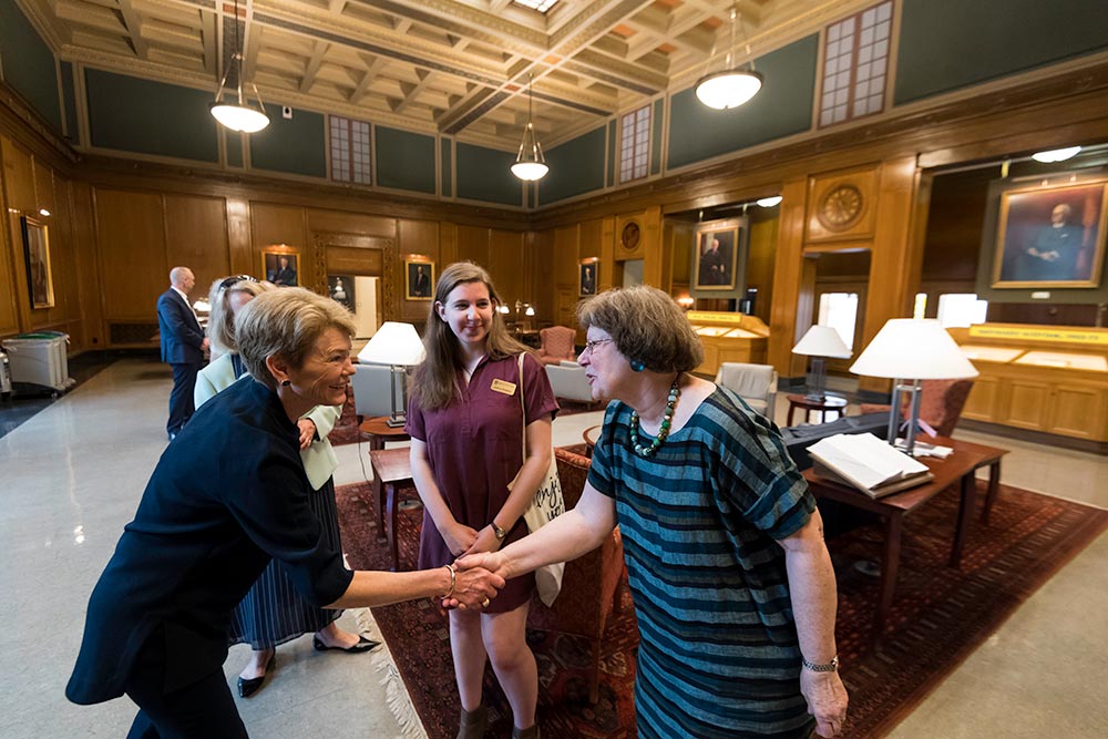 Sarah Mangelsdorf shaking hands with Joan Rubin as others look on in a beautiful library setting.