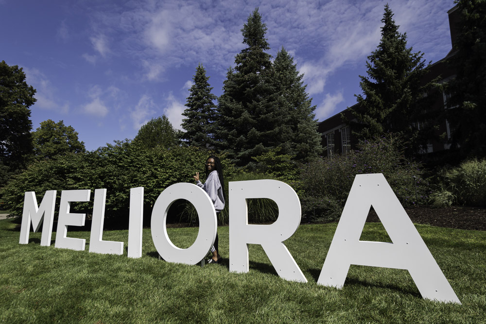 student poses and waves next to large letters that spell MELIORA