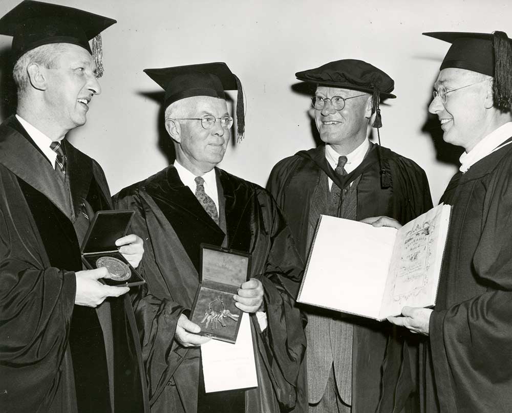four men in academic robes, one holding a ceremonial set of keys, the other holding a charter. 