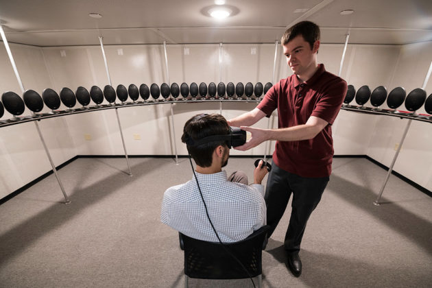 person sitting in a chair wearing a virtual reality headset and surrounded by speakers as another person adjusts the headset