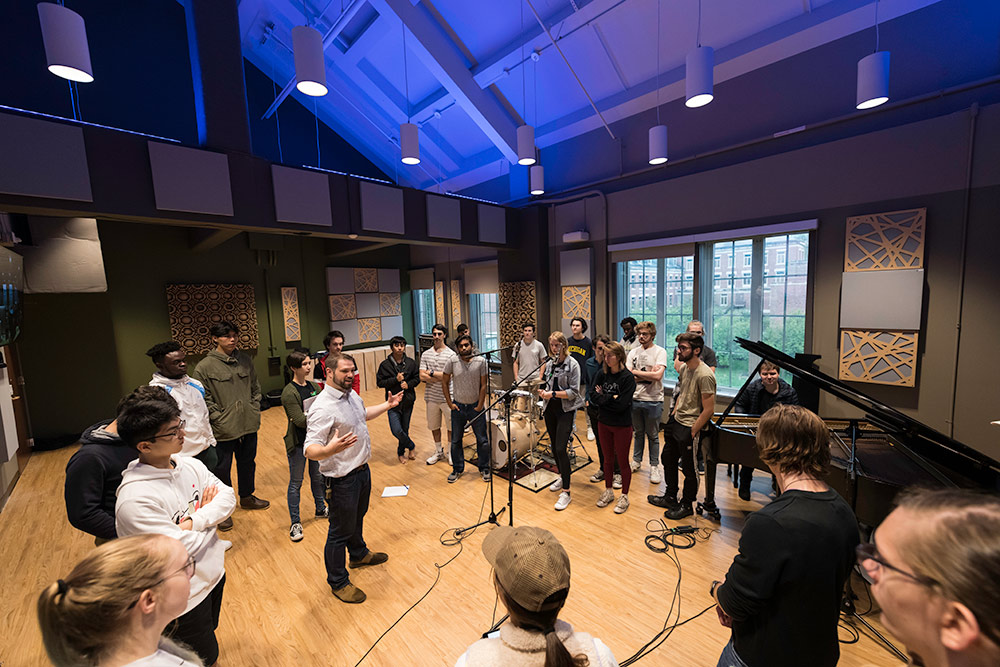 large group of students stands in a circle in recording studio with a piano in the background, listening as a professor teaches. 