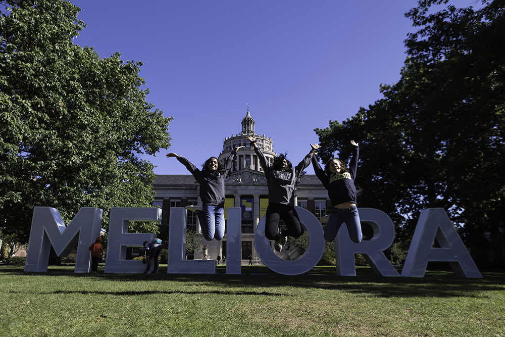 three students jump in front of Meliora letters