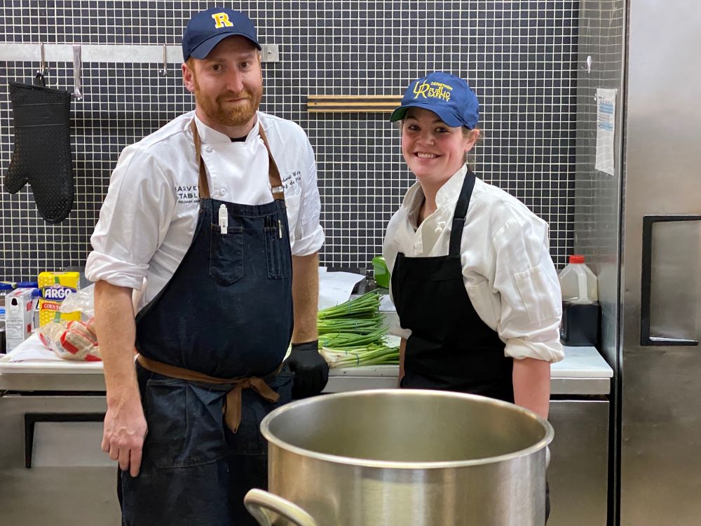 Two people in chef uniforms smile from behind a large pot of boiling water.