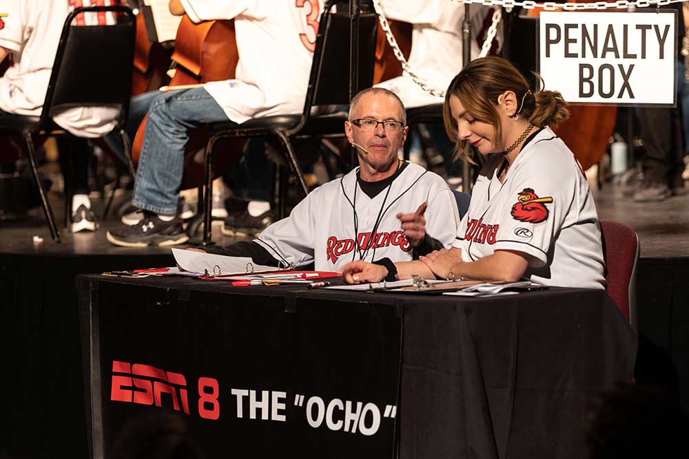 two radio announcer sit at a table in front of a stage, with the words PENALTY BOX and ESPN 8 The Ocho. 