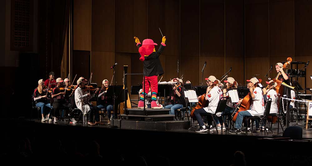 Red Wings baseball mascot stands in front of an orchestra holding a conductors baton.