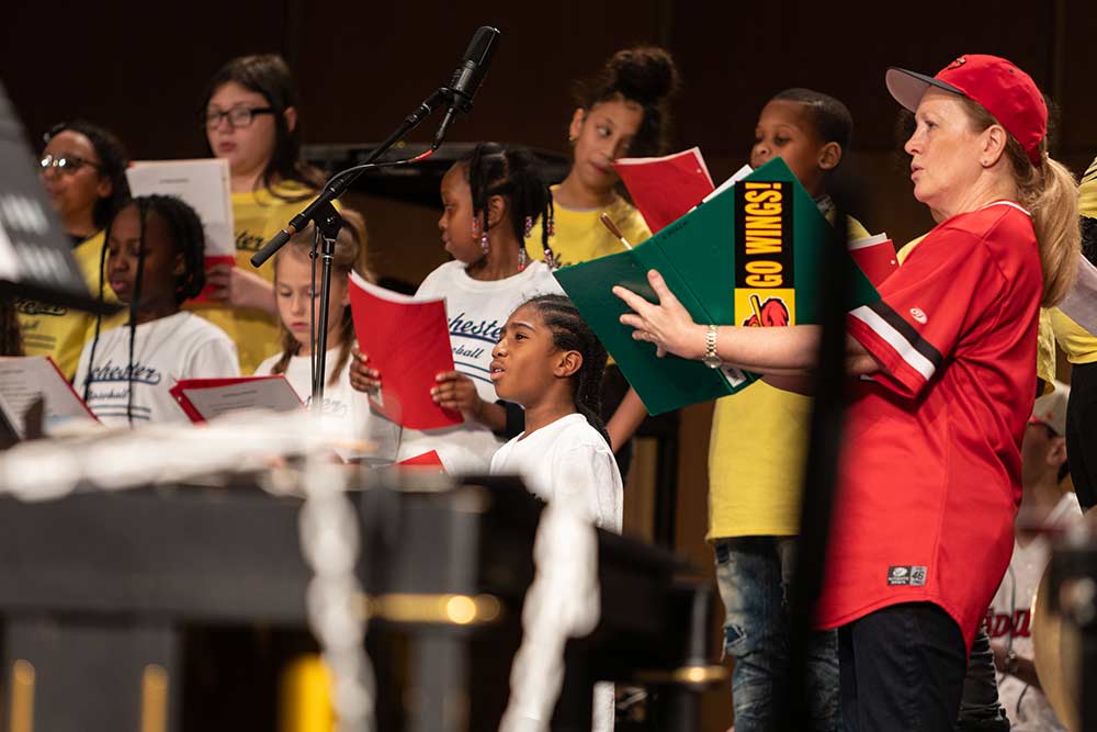 Children's choir perform on stage with choir conductor holding a folder that reads GO WINGS.